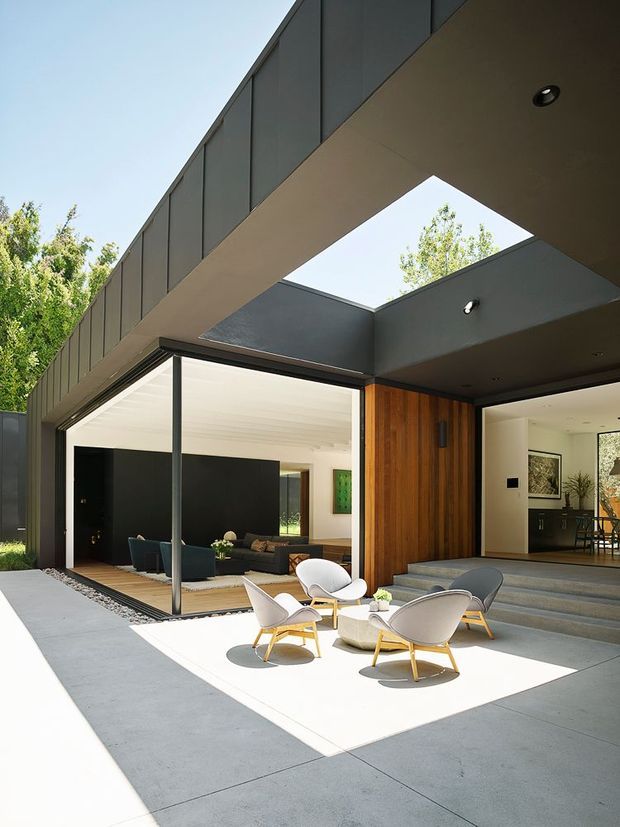 Modern flat-roofed residence with a dramatic cantilevered upper volume and open courtyard terrace. The exterior combines dark metal cladding, warm vertical timber wall cladding, expansive floor-to-ceiling glazing, and a bright concrete-paved outdoor seating area. An open roof courtyard or skylight brings sunlight into the covered entry and entertaining space, while the living room and dining area are visible through the glazing.