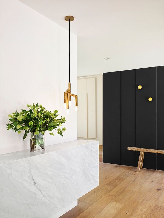 A bright, minimalist entry or kitchen-adjacent interior featuring a large white marble-clad island or console in the foreground, a tall black built-in wall with small brass circular accents, pale timber flooring, white walls, and a suspended brass pendant light. A leafy arrangement in a glass vase and a rustic timber bench add warmth to the otherwise restrained space.