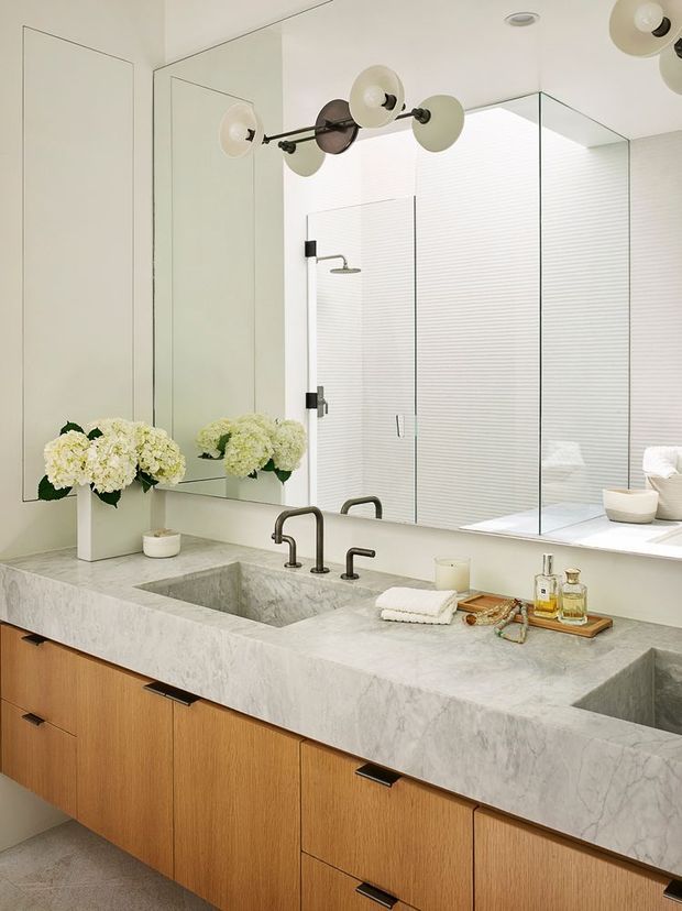 A bright, refined bathroom featuring a large double vanity with a thick pale veined stone benchtop, integrated rectangular basins, warm timber cabinetry, dark metal tapware, and a frameless glass shower enclosure. Full-height mirrors expand the space, while a sculptural multi-globe wall light and floral arrangements add decorative detail.