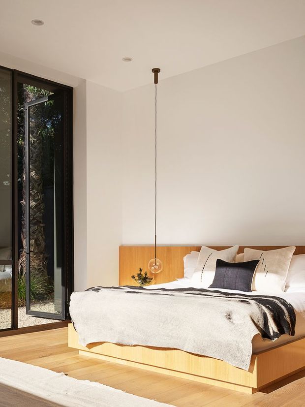 A bright, minimalist bedroom with a low-profile built-in timber bed and matching timber headboard, white bedding, and a black-and-white throw. Full-height dark-framed glazing on the left opens toward greenery, while a simple pendant light hangs above the bedside area. Pale timber flooring and warm natural light create a calm, contemporary atmosphere.