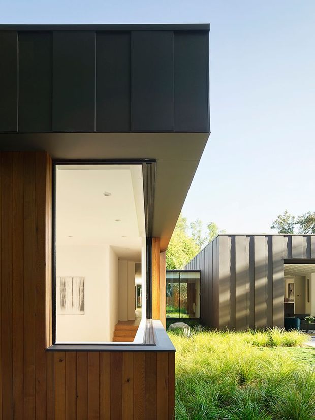 Exterior architectural view of a contemporary low-profile home composed of dark vertical metal cladding, warm timber wall panels, deep roof overhangs, and extensive glazing. The view looks across a landscaped courtyard with ornamental grasses toward a connected dark-clad wing and glazed link. Bright daylight emphasizes the contrast between the dark envelope, timber, and white interior surfaces.