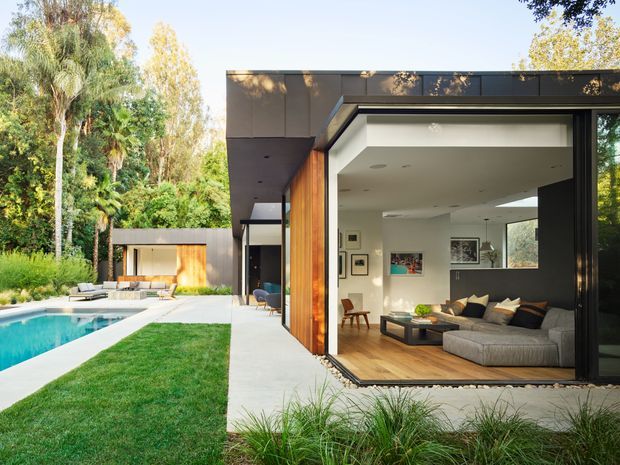 Contemporary single-storey pavilion-style home opening directly onto a landscaped backyard and swimming pool. The image focuses on a spacious living room with fully retractable dark-framed glazing, white walls, warm timber flooring and vertical timber cladding at the opening. A sectional sofa, coffee table and artwork furnish the interior, while the exterior includes a lawn, pool and garden studio-like structure amid mature greenery.