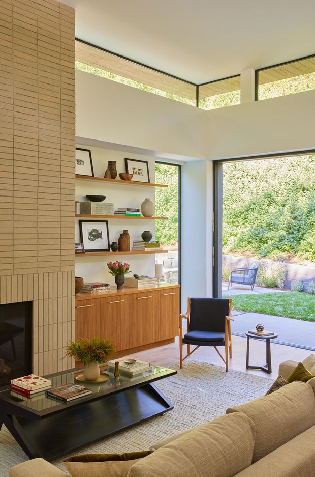 Bright, refined living room with a mid-century-inspired arrangement around a tiled fireplace. The room features a tan brick fireplace surround, warm timber built-in shelving and cabinetry, framed artwork and ceramics, a black glass coffee table, upholstered sofa, wood-framed lounge chair, and large glazed doors opening to a landscaped garden. High clerestory glazing and generous natural light create a strong indoor-outdoor connection.