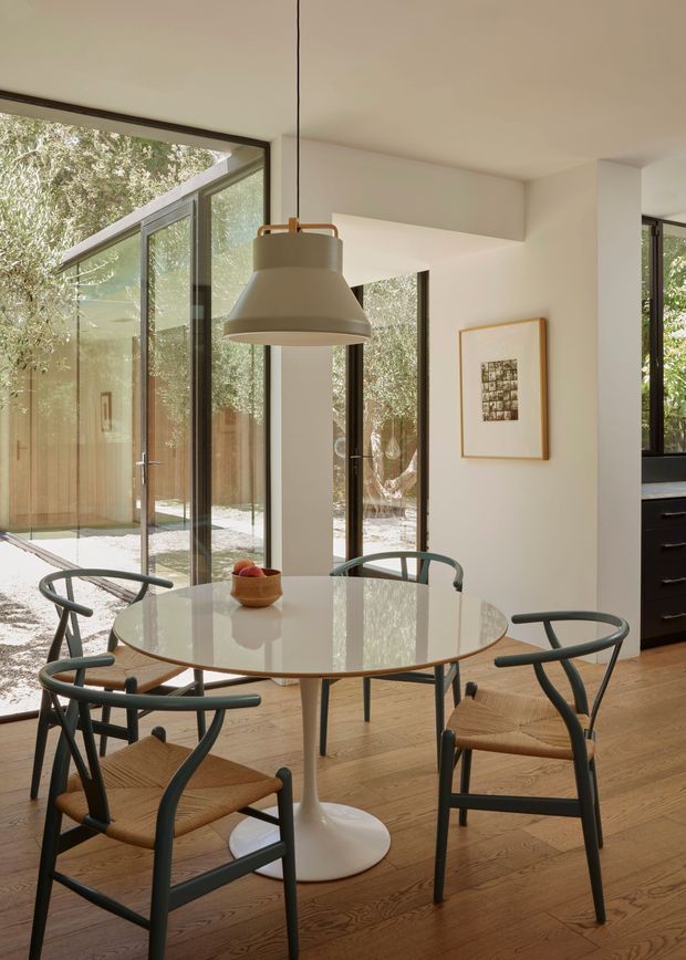 A bright, refined dining area with a round glass-topped pedestal table surrounded by four dark-framed woven dining chairs. A sculptural pendant light hangs above the table. Pale timber flooring, warm white walls, extensive black-framed glazing, and a leafy courtyard visible beyond create a light-filled connection to the outdoors. Dark kitchen cabinetry is partially visible at right, while framed artwork adds a restrained decorative detail.