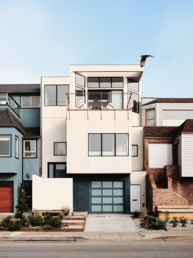 Street-facing photograph of a narrow multi-storey contemporary townhouse or urban residence, with a white rectilinear facade, dark-framed glazing, projecting upper-level volume, glass balcony, and dark garage at ground level. It is tightly flanked by older neighbouring homes, with a small landscaped front setback, steps, and driveway. Clear blue sky and a bird are visible above.
