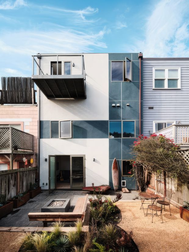 Exterior view of a narrow multi-storey urban residence with a modern geometric facade, blue-grey and white wall panels, large glazed openings, and projecting metal balconies. A landscaped courtyard occupies the foreground with gravel paving, built-in seating, planting, and outdoor chairs. Adjacent timber-clad homes and fencing are visible on both sides under a bright blue sky.