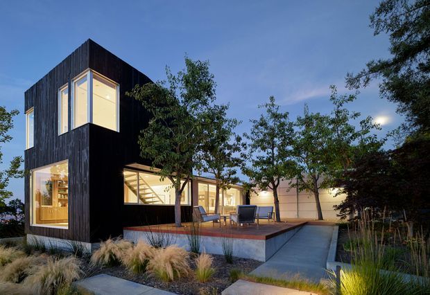 Twilight exterior view of a contemporary multi-level residence with a dramatic black vertically clad upper volume, extensive warm-lit glazing, and a lower wing opening onto a raised timber deck and concrete path. The landscaped approach includes ornamental grasses and mature trees.