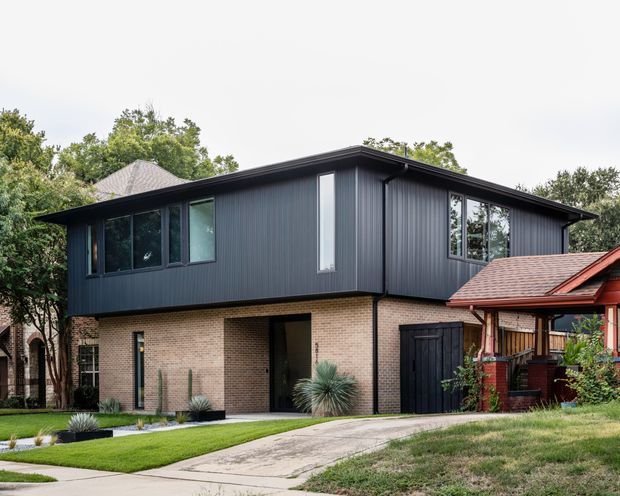 Exterior view of a two-storey contemporary house with dark vertical metal cladding on the upper level, pale brickwork at ground level, a low-pitched roof, and tall narrow windows. A driveway and landscaped front garden lead to the recessed entry and garage, with a neighbouring traditional red-roofed structure visible at the right.