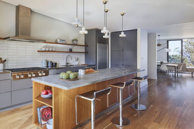A bright, open-plan contemporary kitchen with a large island clad in warm timber and topped with grey stone. The kitchen features matte grey cabinetry, a stainless-steel rangehood and cooker, white rectangular tiled splashback, open timber shelving, and four modern bar stools. Pendant lights hang above the island, while a dining and living area is visible beyond, with dark timber flooring continuing throughout.