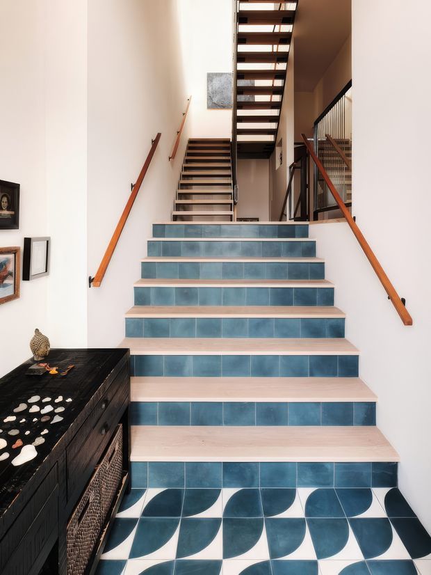 Bright interior stairway with a broad staircase finished in blue tile risers and a patterned blue-and-white tiled landing. Pale timber or light-toned stair treads contrast with the blue surfaces. Warm timber handrails run along white walls, while a dark metal balustrade and open-riser staircase continue upward. A dark timber console with decorative objects sits beside the stairs, and framed artwork is mounted on the wall.