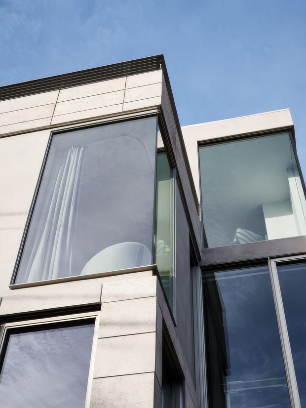 Upward-looking architectural photograph of a modern multi-storey building facade with large floor-to-ceiling glazing, dark metal window frames, pale stone or concrete cladding, and strong projecting geometric volumes against a blue sky.