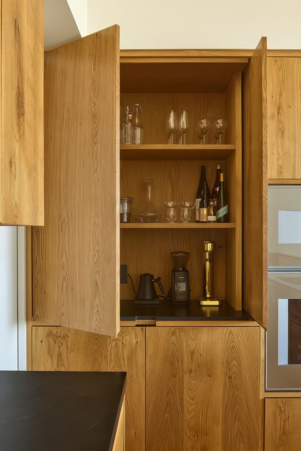Close-up view of a built-in timber kitchen cabinet with open doors, displaying glassware, bottles, a coffee grinder, kettle, and other beverage accessories. The cabinetry has a strong vertical wood grain and warm natural finish, with a dark benchtop visible in the foreground and partial integrated appliances at right.
