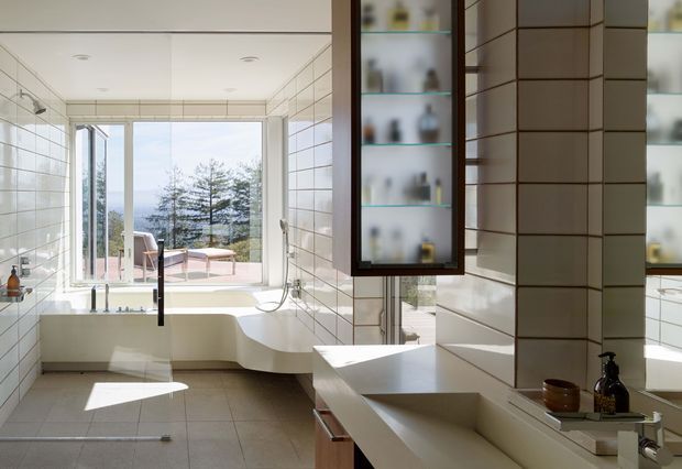 Bright contemporary bathroom with a freestanding bathtub positioned beside large floor-to-ceiling glazing, a separate vanity with an integrated rectangular basin, wall-to-wall white rectangular tiles, recessed ceiling lights, and a dark-framed mirrored or glass-fronted storage cabinet. The room opens visually to an outdoor terrace and trees beyond.