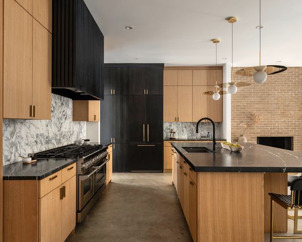 A sophisticated open-plan kitchen with a large central island, warm timber cabinetry, dark stained timber and black accents, veined marble-look splashbacks, and dark stone benchtops. A black-framed fireplace and exposed red-brick wall are visible in the adjoining area. The space features sculptural globe pendant lights, recessed ceiling lights, integrated appliances, and a refined contemporary industrial character.