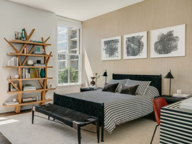 A bright, refined bedroom with a black upholstered bed dressed in a bold black-and-white patterned coverlet and pillows. A woven black bench sits at the foot of the bed, while an angular timber bookshelf displays books and objects beside a tall window. The room features a textured beige wall with three framed abstract artworks, black bedside lamps, and a striped black-and-white bedside cabinet with a red chair partially visible. The overall design is contemporary with graphic monochrome accents and warm timber detailing.