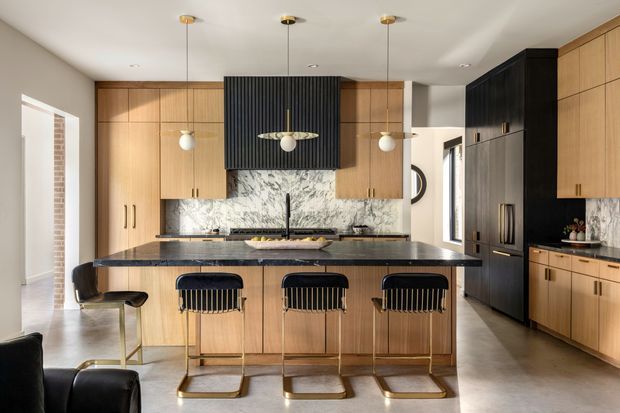 A refined contemporary kitchen centered on a large island with seating for four. The room features light timber cabinetry, dark charcoal-black tall cabinetry and a vertically fluted rangehood enclosure, a dramatic veined stone splashback, dark stone island benchtop, polished concrete-look flooring, and brass-toned pendant and stool details. The open-plan space is bright and carefully balanced with warm timber and dark contrasting elements.