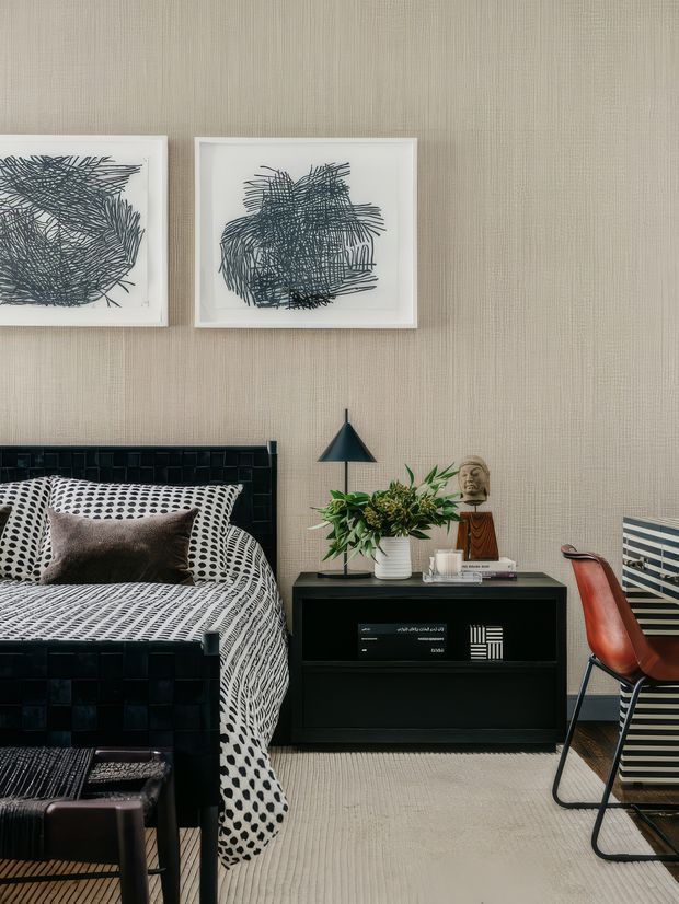 A stylish bedroom with a black tiled or panelled bed frame, patterned black-and-white bedding, and a dark bedside cabinet. The room features textured beige wall covering, abstract monochrome artwork, a black cone-shaped table lamp, indoor greenery, sculptural decor, and a red leather desk chair beside a striped desk or cabinet. The look is graphic, curated, and contemporary with mid-century influences.
