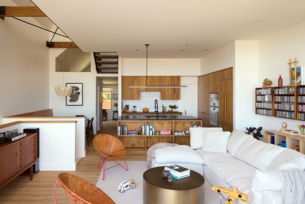 Open-plan contemporary living, dining and kitchen interior with warm timber joinery, exposed timber ceiling beams, a compact island, integrated shelving, white walls, and a large white sectional sofa. A stairway and partial upper-level structure are visible, along with a wall-mounted record or book collection and eclectic furnishings.