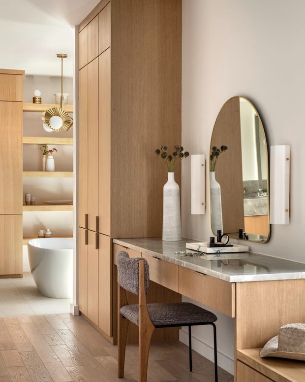 A warm, refined bathroom vanity and dressing area with custom light- to mid-toned timber cabinetry, a pale stone benchtop, an arched wall mirror, wall sconces, and a timber-framed upholstered chair. The space opens toward a partially visible freestanding bathtub, built-in shelving, and additional bathroom fittings. Timber flooring continues through the room, creating a cohesive contemporary interior.