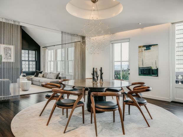 Refined open-plan dining area with a round wood-and-glass dining table surrounded by sculptural mid-century style wooden chairs. The room features dark timber flooring, a large pale circular rug, white walls and trim, sheer floor-to-ceiling curtains, tall divided-light windows, a contemporary sculptural chandelier, and an adjacent lounge area. A black geometric fireplace or architectural feature is visible to the left.