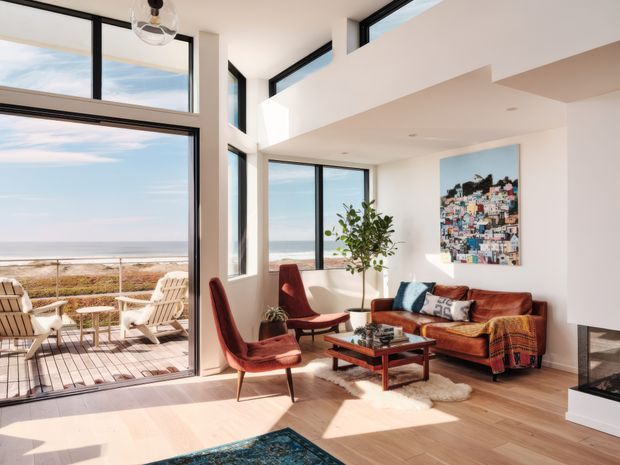 Bright, double-height coastal living room with expansive black-framed glazing overlooking the ocean. The room features a warm timber floor, white walls, a brown leather sofa, mid-century-style lounge chairs, a low timber coffee table, a contemporary fireplace, and a large colourful artwork. An adjoining outdoor deck is visible through the open glazing.