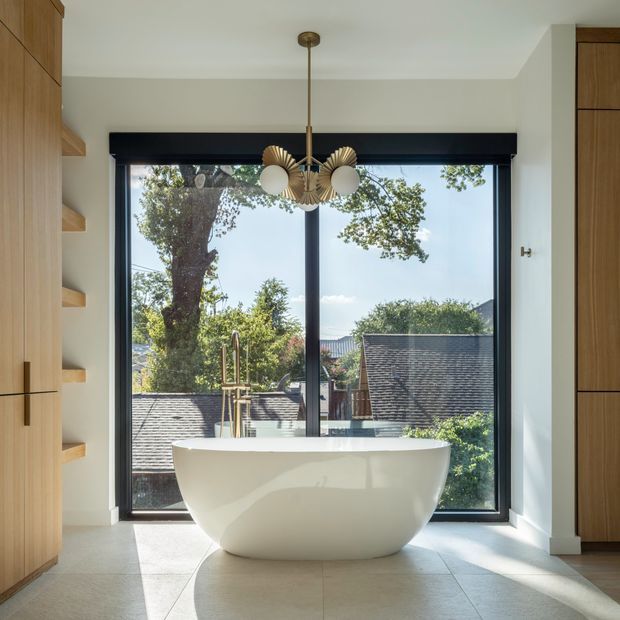 A bright, refined bathroom centred on a freestanding white oval bathtub positioned in front of full-height black-framed glazing. Warm timber built-in cabinetry and open shelving flank the room, while a sculptural brass-and-white globe pendant hangs above. Large pale stone or porcelain floor tiles and a leafy residential outlook create an airy, contemporary setting.