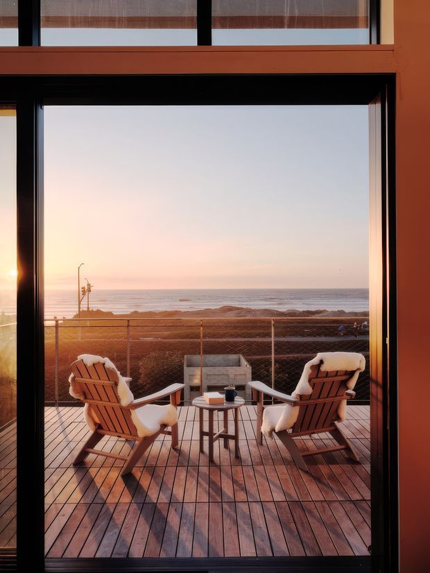 View from inside through a large black-framed glazed opening onto a timber deck overlooking the ocean at sunset. Two cushioned timber lounge chairs and a small side table form an outdoor seating area, with a glass balustrade and coastal beach beyond.