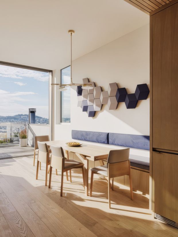 A bright, contemporary dining area with a light timber dining table and chairs, built-in timber cabinetry and banquette seating upholstered in muted blue. Geometric hexagonal wall art adds a sculptural accent above the banquette. Large corner glazing opens to expansive urban and hillside views, while a linear pendant light hangs above the table. The room has pale timber flooring, warm natural light, and a partially slatted timber ceiling detail.