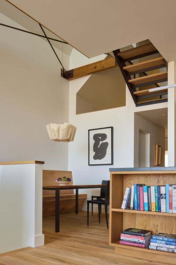 A warm, contemporary open-plan interior featuring a dining nook, timber flooring, built-in timber shelving, and a dramatic open-riser staircase with dark structural framing. A suspended sculptural pendant hangs above the dining area, while exposed timber beams and high ceilings create a spacious, loft-like atmosphere. Neutral plaster walls, black-framed artwork, and carefully integrated joinery give the space a refined modern character.
