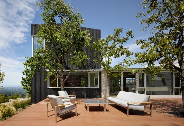 A modern multi-level home viewed from a broad timber deck, with outdoor lounge furniture arranged around a low coffee table. The house has dark vertical cladding, extensive glazing, a flat or low-profile roofline, and a partially covered terrace. Mature trees frame the view, with a distant landscape visible beyond the property.