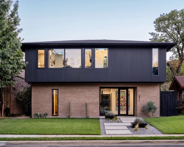 Front exterior view of a contemporary two-storey residence. The lower level is clad in light brown brick, while the upper level projects outward and is finished in dark vertically ribbed cladding. Large rectangular windows, a glazed entry door with decorative coloured glass, a paved stepping-stone path, gravel landscaping, lawn and established trees are visible.