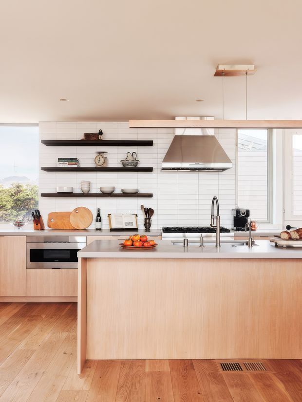Bright contemporary kitchen with a large timber-fronted island, integrated cooktop and sink, white horizontal wall tiling, open dark shelves, stainless-steel rangehood, pale timber cabinetry, and timber flooring. Large windows bring in daylight and views of greenery.