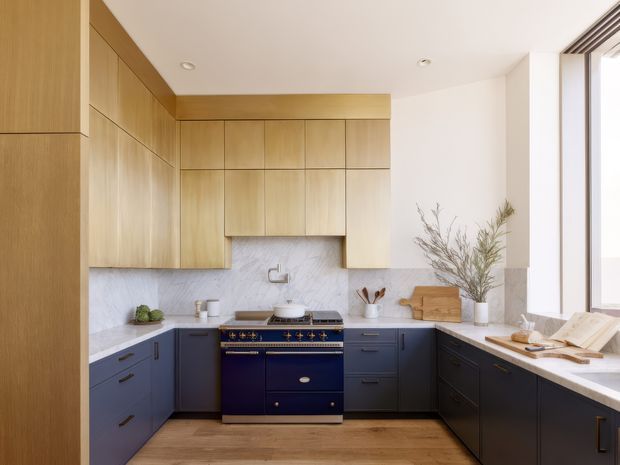 A bright, refined contemporary kitchen with a U-shaped layout. The room features full-height light timber cabinetry, deep navy-blue lower cabinets, a large navy range cooker, pale veined stone benchtops and backsplash, and warm timber flooring. A tall window brings in abundant natural light, while restrained styling includes ceramics, a cutting board, cookbooks, and dried branches.