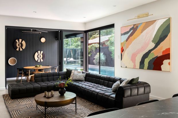 A stylish contemporary living room with a large black modular sectional sofa arranged around a round coffee table. The room features a dramatic black fluted feature wall with decorative circular wall art, expansive black-framed glazing overlooking a pool courtyard, a large abstract artwork with a picture light, recessed ceiling lights, and a patterned area rug. A dark stone kitchen island or benchtop is visible in the foreground.