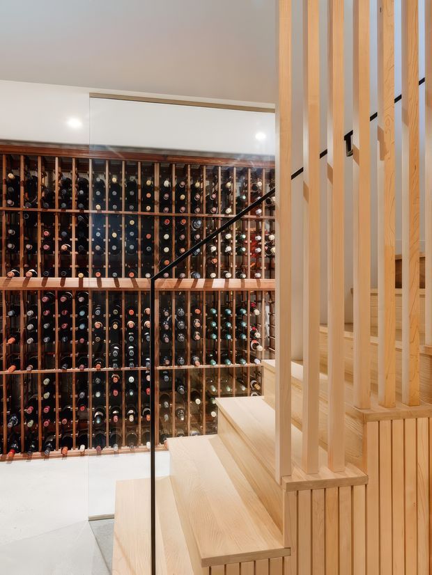 Interior view of a refined wine storage area beneath a light timber staircase. A large glass-fronted wine cellar wall is densely lined with horizontally stored bottles, while pale timber stair treads, vertical balustrade slats, and panelled joinery occupy the foreground. The space has warm integrated lighting, a clean contemporary fit-out, and a strong timber-and-glass palette.