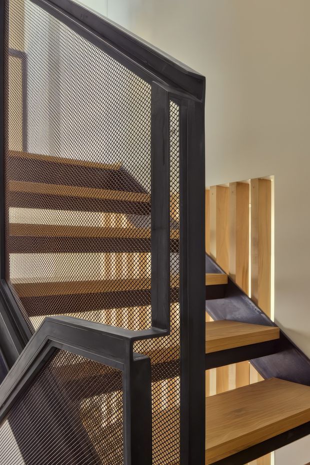 Interior architectural photograph of a contemporary stairway with open-riser timber treads, dark steel framing, and black expanded-metal mesh balustrades. Warm timber-lined wall openings and a stair flight are visible behind the prominent screen, creating a layered, sculptural circulation space.