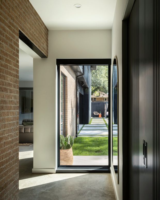 A contemporary residential entryway viewed from inside, with a full-height glazed door opening to a landscaped courtyard. The interior combines an exposed red brick wall, smooth off-white plaster walls, a polished grey concrete floor, and dark vertically ribbed built-in cabinetry. A living room is partially visible through the opening on the left, while the courtyard beyond contains lawn, concrete stepping slabs, brick walls, and dark cladding.