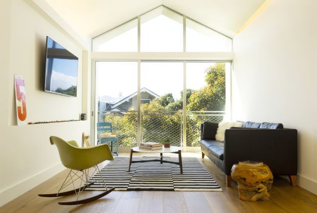 Bright, compact living room with a dramatic gabled, floor-to-ceiling glazed wall overlooking neighbouring rooftops and trees. The room has pale walls, warm timber flooring, a black sofa, lime-green sculptural rocking chair, low timber coffee table, striped area rug, wall-mounted television, and a sculptural yellow side table. The space has a clean mid-century-influenced contemporary character and strong natural light.