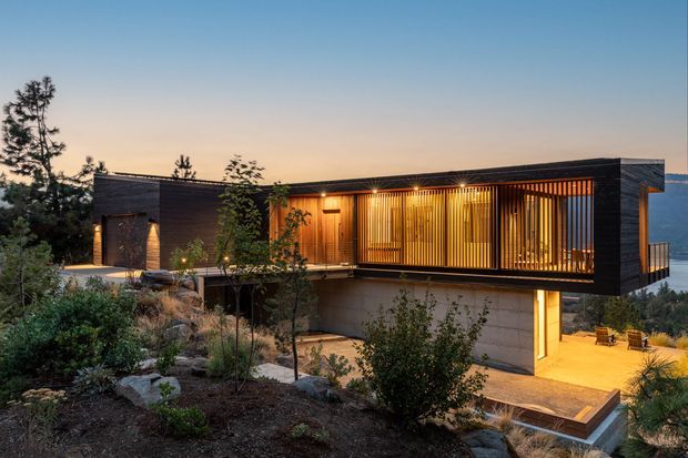 A modern, low-profile multi-level house set into a landscaped sloping site at dusk. The dark horizontal exterior is contrasted by warm timber screening and extensive glazed openings, with a sheltered terrace and illuminated lower-level outdoor area. Mountainous terrain is visible in the distance.