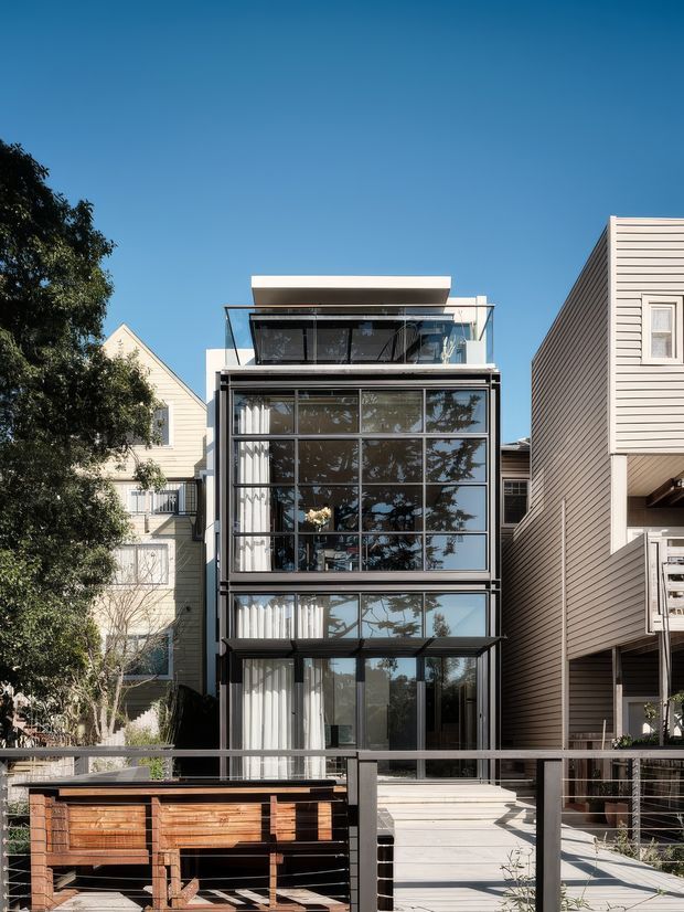 A narrow multi-storey contemporary townhouse facade viewed from the street or front terrace. The building has extensive floor-to-ceiling glazing in dark metal frames, projecting glass-fronted levels, a flat roof, and horizontal cladding. A timber outdoor structure and deck railing occupy the foreground, with neighbouring residential buildings closely framing the property.