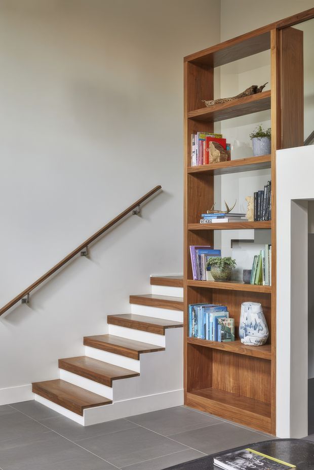 Interior view of a modern stairway with floating timber treads, a dark timber handrail, and white walls. A full-height built-in timber shelving unit runs alongside the stairs, displaying books and decorative objects. Large grey floor tiles and clean-lined detailing create a restrained, contemporary feel.
