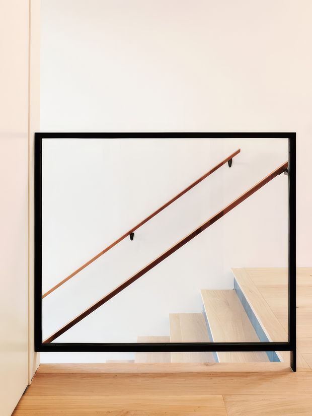 Minimalist interior view of a stairwell through a black-framed glazed partition or balustrade. Pale timber stair treads and flooring contrast with white walls, while slim warm timber handrails run diagonally along the staircase. The composition is clean, bright, and architectural, with a strong geometric emphasis.