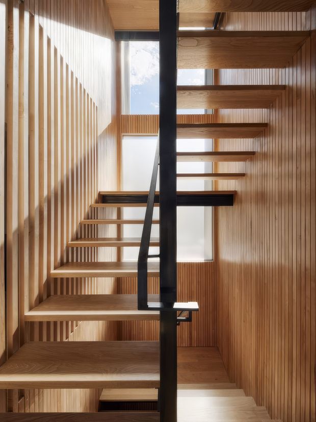 A tall, light-filled interior stairway with open-riser timber stairs, slim black metal handrails and a central dark structural element. Vertical timber slat wall panelling wraps the stairwell, while clerestory-like windows and roof glazing bring in daylight. The composition emphasizes the stair void and layered landings.