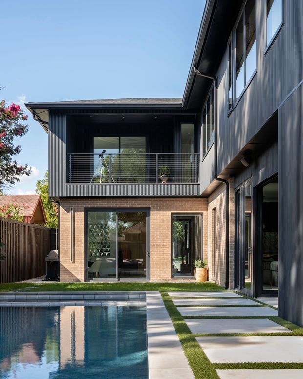 A contemporary multi-level residential courtyard viewed from beside a swimming pool. The house combines dark vertically ribbed cladding, exposed warm brickwork, extensive glazing, and a projecting upper-level balcony with horizontal metal balustrades. Large pale concrete pavers separated by narrow strips of grass lead along the pool edge toward the glazed ground-floor spaces. The composition is bright and architectural, with strong reflections in the pool and a sheltered courtyard setting.