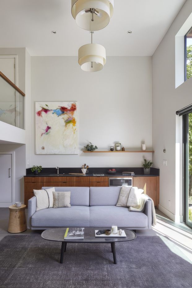 Bright, double-height open-plan living room with a compact integrated kitchen along the rear wall. The room features a pale grey upholstered sofa, dark textured rug, oval coffee table, timber slatted cabinetry, dark benchtop, open timber shelf, large abstract artwork, pendant drum lights, and tall glazing bringing in strong daylight. A partial upper-level landing or mezzanine is visible at left.