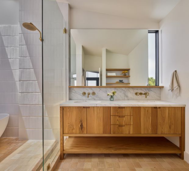 Bright, refined bathroom featuring a long floating timber vanity with integrated open shelving, a white marble benchtop and matching marble splashback, brass wall-mounted tapware, a large mirror, and a glass-enclosed shower lined with textured white tiles. Warm beige tiled flooring and soft neutral walls create a calm, elevated interior.