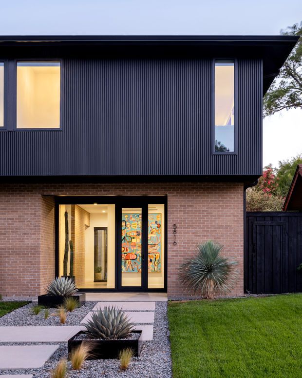 Front exterior view of a contemporary two-storey residence. The upper level is clad in dark vertically ribbed metal or composite-style panels beneath a broad flat roof, while the lower level uses warm red brick. Large black-framed windows and a glazed entry door with colourful decorative panels face a landscaped front path featuring gravel, concrete stepping slabs, ornamental grasses and sculptural agave plants. A dark timber boundary fence and neighbouring roofline are visible at the side.
