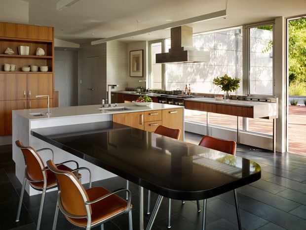 A spacious, light-filled contemporary kitchen and dining area with an open-plan layout. The room features a large glossy black dining table with leather-and-metal chairs, a white waterfall-edge island with integrated seating, warm timber cabinetry, dark tiled flooring, stainless-steel appliances and rangehood, and extensive floor-to-ceiling glazing opening onto a timber deck and landscaped exterior. Clean horizontal lines and abundant natural light create a refined mid-century-influenced atmosphere.