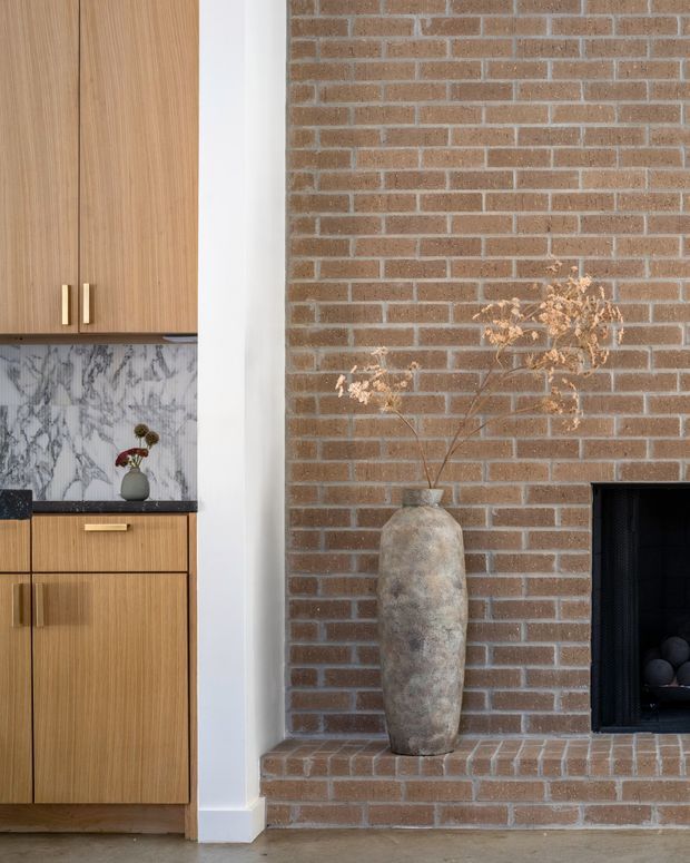 Close-up interior view showing a built-in timber cabinet and drawers with brass pulls beside a substantial exposed brick fireplace. The cabinetry includes a dramatic grey-white marble backsplash and dark benchtop, while a tall ceramic vase sits on the brick hearth. The composition is warm, textural, and contemporary with strong natural materials and clean detailing.