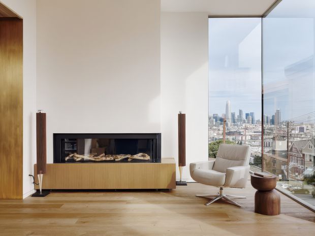 A refined contemporary living room with a full-height corner window overlooking a dense city skyline. The room features a long built-in fireplace with black framing and a warm wood media cabinet, pale plaster walls, wide timber flooring, sculptural wood speakers, and a cream lounge chair with a matching small wood side table. The expansive glazing and restrained furnishings create a bright, airy, minimalist atmosphere.